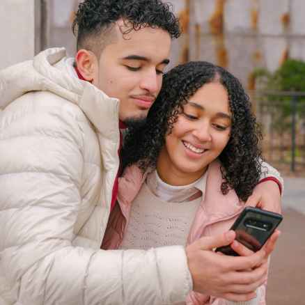 smiling young ethnic couple hugging and using smartphone on street